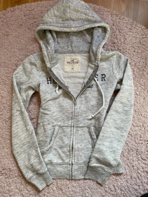 Grå zip hoodie från Hollister XS - Ljusgrå hoodie från Hollister med dragkedja, broderad text på bröstet och två fickor framtill. Tröjan har huva med dragsko och ribbade muddar vid ärmar och nederkant. Perfekt för en avslappnad och sportig stil.
