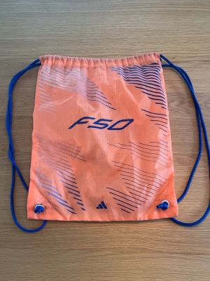 Orange ryggsäck från Adidas F50 - Cool orange ryggsäck från Adidas F50 med blå snören och grafiska detaljer i blått. Perfekt för att bära träningskläder eller småsaker. Väskan är lätt och har ett sportigt utseende med F50-logga på båda sidor.