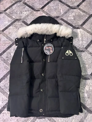 Svart dunjacka från Moose Knuckles - Säljer en svart dunjacka från Moose Knuckles med avtagbar huva och vit fuskpäls. Jackan har silverfärgade dragkedjor, tryckknappar och den ikoniska logotypen på ärmen. Perfekt för kalla vinterdagar och riktigt snygg streetstil.