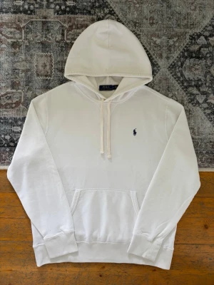 Vit Polo Ralph Lauren Hoodie - Storlek L - Hej! 👋 Säljer en klassisk vit Polo Ralph Lauren hoodie i storlek L med marinblå pony-logga. 🐎✨ Tröjan är i fint skick överlag men har blivit något nopprig. Det finns en liten prick på ena ärmen (se sista bilderna) samt eventuellt någon mer minimal prick, inget som märks vid användning. 100% äkta med originaltvättlappar och en fullt fungerande QR-kod på lappen för enkel verifiering ✅ Perfekt lyxigt basplagg! Fraktar snabbt! 💬 💯 🚛📦 