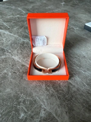  armband med guldspänne - Snyggt armband från H i svart med guldfärgat H-spänne. Kommer i original orange ask och medföljer äkthetskort och dustbag. Armbandet har en stilren och lyxig design i metall och emalj, perfekt för att lyfta din outfit.