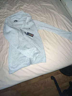 Grå hoodie från Polo Ralph Lauren - Snygg ljusgrå hoodie från Polo Ralph Lauren med dragkedja framtill och klassisk huva med dragsko. Liten broderad logga på bröstet och mjukt material som känns skönt mot huden. Perfekt för en avslappnad och stilren look.