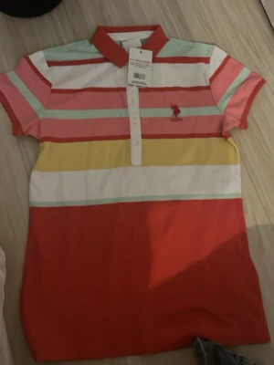 Randig pikétröja från U.S. Polo Assn. - Färgglad pikétröja från U.S. Polo Assn. med breda ränder i rött, rosa, gult, mintgrönt och vitt. Klassisk krage och knappar framtill. Broderad logga på bröstet. Perfekt för en avslappnad och sportig look.