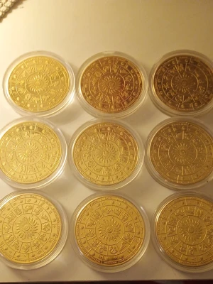 Guldiga zodiakmynt i plastfodral - Samling med nio runda guldiga mynt med zodiakens tecken och solmotiv, tillverkade i metall och förvarade i genomskinliga plastfodral. Perfekt för dig som gillar astrologi eller vill ha en unik dekoration till rummet.