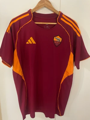AS Roma matchtröja Adidas vinröd - Säljer en vinröd AS Roma matchtröja från Adidas med orange detaljer och klubbmärke på bröstet. Tröjan har korta ärmar, rund hals och är tillverkad i ett lätt, andningsbart material. Perfekt för fotbollsträning eller att bära till vardags.