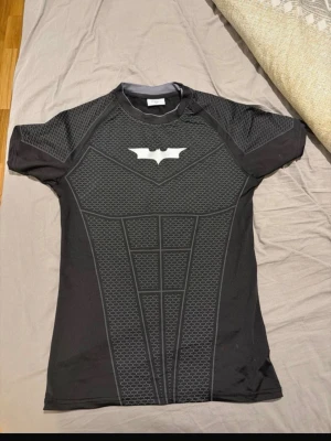 Batman comprision t-shirt från YoungLA - Snygg svart Batman cosplay t-shirt från YoungLA i storlek M. Tröjan har tryckt Batman-logga på bröstet och detaljer som liknar Batmans dräkt. Tillverkad i stretchigt, mjukt material som passar perfekt till maskerad eller cosplay.