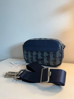 Lacoste Axelväska Mörkblå  - Snygg mörkblå axelväska från Lacoste med monogram mönster i ljusblått. Väskan har justerbar axelrem i textil och detaljer i metall. Praktisk storlek för det viktigaste och stilren design med Lacoste-logga på framsidan.