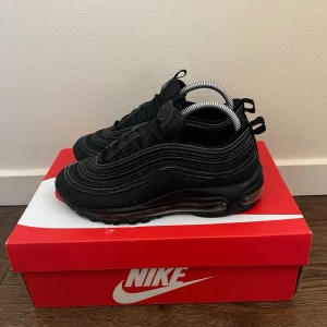 Nike Air Max 97 helsvarta sneakers - Nike Air Max 97 sneakers i helsvart med ikoniska vågformade linjer och synlig Air-enhet i sulan. Ovandelen är i mesh och syntetmaterial för en sportig look. Skorna har reflekterande detaljer och klassisk snörning. Perfekta för dig som gillar streetwear och stilren design.