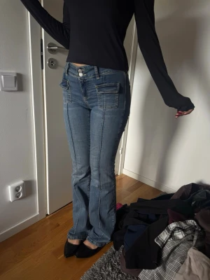 Blå bootcut jeans med fickor - Säljer ett par ljusblå bootcut jeans med markerade sömmar och stora fickor fram. Jeansen har normal midja och klassisk femficksdesign. Tillverkade i denim med lätt stretch för skön passform. Perfekta för en avslappnad och trendig look.