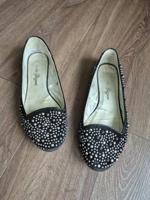 Svarta ballerinaskor med pärlor - Snygga svarta ballerinaskor från Salvatore Ferragamo, dekorerade med mängder av nitar över hela skon. Skorna har rundad tå och är tillverkade i mocka för en lyxig känsla. Perfekta för dig som vill sticka ut med en unik och trendig look.