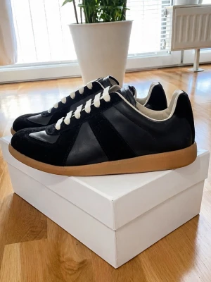 Maison Margiela Gats - Stilrena svarta sneakers från Maison Margiela med ovandel i läder och mocka, vita skosnören och beige gummisula. Klassisk low-top siluett med retrovibbar och diskreta detaljer. Perfekta för dig som gillar minimalistisk och tidlös design.