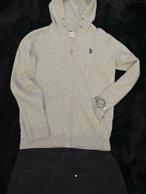 Grå zip hoodie från POLO ASSN - Har ett litet omärkbar fläck (På sista bilden) Snygg grå hoodie med dragkedja från U.S POLO ASSN. Klassisk huva med dragsko, två fickor fram och diskret logga på bröstet samt flaggdetalj vid ärmslutet. Perfekt till chill vardagsstil och enkel att matcha med jeans eller joggers.