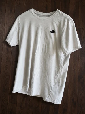 Vit Nike t-shirt med logga - Klassisk vit t-shirt från Nike med svart broderad logga på bröstet. T-shirten har rund halsringning och korta ärmar. Tillverkad i mjuk bomull för en skön och avslappnad stil.