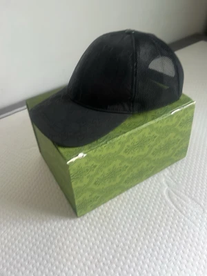 Svart meshkeps från Gucci - Säljer en svart keps från Gucci med meshpaneler på sidorna och justerbar snapback baktill. Kepsen har diskret GG-mönster på framsidan och logotyp på baksidan. Perfekt för dig som vill ha en stilren och exklusiv accessoar.
