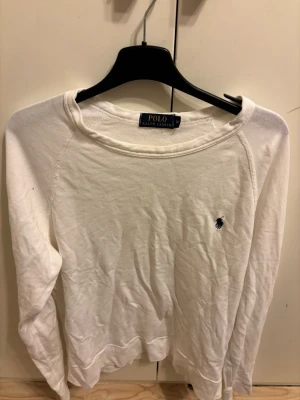Vit tröja från Polo Ralph Lauren - Klassisk vit långärmad tröja från Polo Ralph Lauren med rund halsringning och den ikoniska lilla marinblå logon broderad på bröstet. Tröjan har en enkel och stilren design, perfekt för en clean look. Tillverkad i mjukt bomullsmaterial.