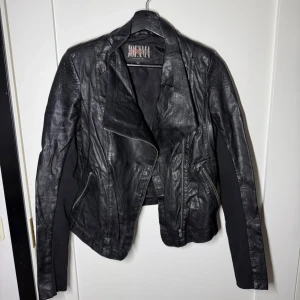 Svart skinnjacka från Jofama - Snygg svart skinnjacka från Jofama med asymmetrisk dragkedja och coola detaljer. Jackan har två snedställda fickor med dragkedja och en modern, figurnära passform. Perfekt för dig som vill ha en edgy look.