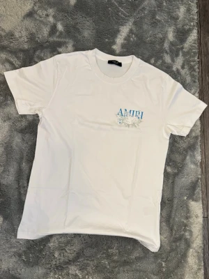 Vit AMIRI t-shirt Storlek M - Vit t-shirt från AMIRI med blå logga och blommigt tryck både fram och bak. Klassisk rund hals och korta ärmar. Perfekt för dig som gillar stilrena plagg med en twist. Storlek M