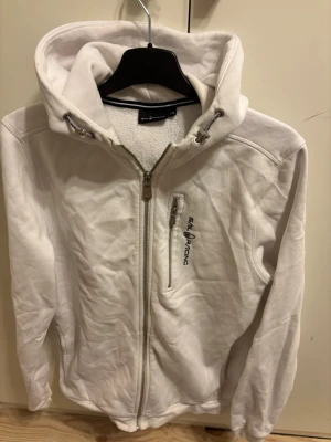 Vit hoodie från Sail Racing - Vit hoodie från Sail Racing med dragkedja framtill och bröstficka med logga. Huva med dragsko och broderad detalj bak. Mjuk fleecekänsla och långa ärmar. Perfekt för dig som gillar stilren och sportig look.