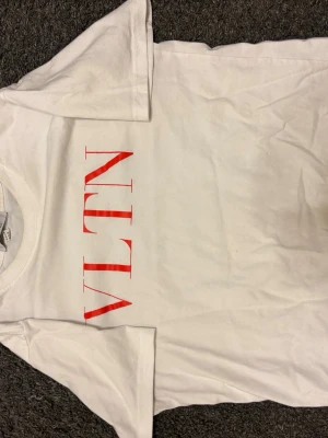 Vit t-shirt med röd VLTN-tryck - Vit t-shirt från Valentino med stort rött VLTN-tryck på framsidan. Klassisk rund hals och korta ärmar, perfekt för en clean och stilren look. Materialet är mjuk bomull som känns skönt mot huden.