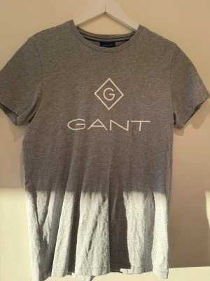 Grå GANT t-shirt med logga - Snygg grå t-shirt från GANT i mjuk bomull. Klassisk passform med rund hals och korta ärmar. Stor vit GANT-logga framtill som ger en clean och stilren look. Perfekt till jeans eller shorts för en enkel vardagsstil.