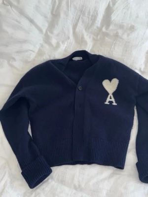 Mörkblå cardigan från Ami Paris - Snygg mörkblå cardigan från Ami Paris med vitt hjärta och bokstaven A på bröstet. Koftan har v-ringning, ribbade muddar och knäppning framtill. Perfekt för lager på lager och ger en trendig look.