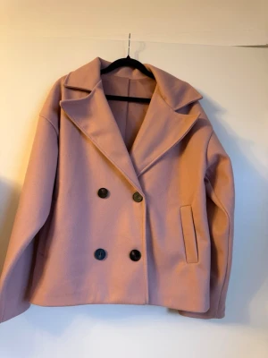 Ljusrosa dubbelknäppt rock - Snygg ljusrosa rock med dubbelknäppning och stora svarta knappar. Klassisk krage och raka fickor framtill. Kortare modell med rak passform, perfekt för att lyfta din outfit.