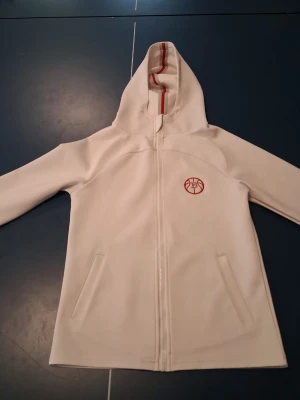Vit hoodie med röd detalj och dragkedja - Snygg vit hoodie med hel dragkedja och röda detaljer på huvan. Märket 2HS är broderat på bröstet. Hoodien har två fickor framtill och är tillverkad i ett mjukt material som känns skönt mot huden. Perfekt för en avslappnad och sportig stil.