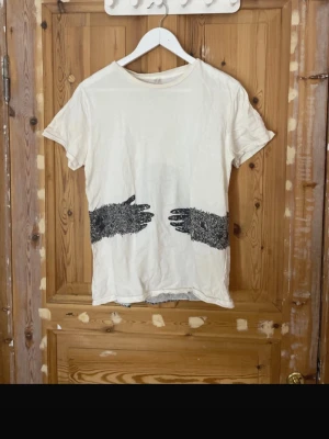 Vit t-shirt med konstnärligt tryck - Unik vit t-shirt med svart konstnärligt tryck av två händer framtill och ett abstrakt mönster på ryggen. T-shirten har rund hals och korta ärmar. Materialet känns mjukt och passar perfekt för en avslappnad stil. Finns missfärgningar men inget som syns vid användning 