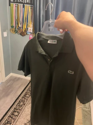 Svart pikétröja från Lacoste - Klassisk svart pikétröja från Lacoste med vit broderad logga på bröstet. Tröjan har krage, knappar framtill och korta ärmar. Perfekt till en avslappnad men ändå stilren look.