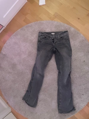 Grå raka jeans med fickdetaljer - Säljer ett par grå jeans med rak passform och snygga fickor med lock och knapp bak. Jeansen har klassisk femficksdesign och är i ett tvättat denimtyg. Perfekta för en avslappnad och cool stil.