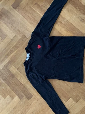 Svart långärmad tröja Comme des Garçons - Svart långärmad topp från Comme des Garçons Play med det ikoniska röda hjärtat broderat på bröstet. Tillverkad i mjuk bomull med rund halsringning. Perfekt för en clean och stilren look. Medium men passar som S