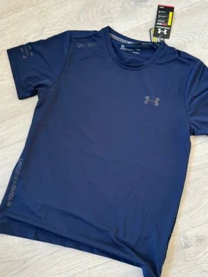 Blå träningströja från Under Armour - Snygg mörkblå t-shirt från Under Armour med diskret logga på bröstet och tryck på ärmen. Tillverkad i ett lätt och snabbtorkande funktionsmaterial som passar perfekt till träning och sportaktiviteter. Klassisk rund hals och korta ärmar.