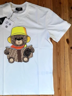 Vit Fendi t-shirt med nalletryck - Vit t-shirt från Fendi med ett stort tryck av en nallebjörn som läser en bok med Fendi-logga. Nallen har en gul hatt och orange scarf. Klassisk passform och rund halsringning. Perfekt för dig som gillar statement-plagg och coola designer.