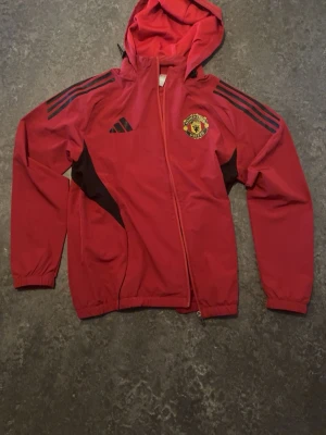 Manchester United windbreaker tracksuit - Säljer ett Manchester United träningsset från Adidas. Jackan är röd med svarta detaljer, huva och klubbmärke på bröstet. Byxorna är svarta med röda ränder och klubbmärke. Perfekt för fotbollsträning eller chill. 