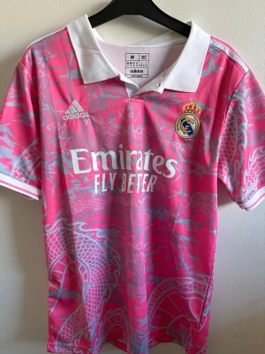 Real Madrid rosa tröja  Adidas - Säljer en rosa Real Madrid matchtröja från Adidas med vitt krage och detaljer. Tröjan har ett stort drakmönster i silver över hela plagget, klubbmärke på bröstet och sponsortryck framtill. Om det e några frågor så va inte rädd att fråga