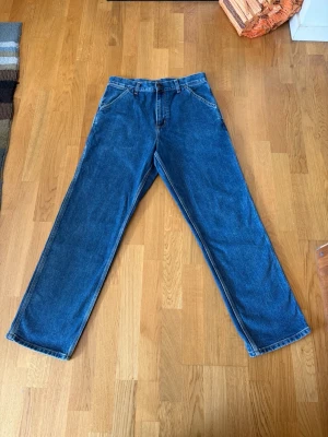 Blå raka carpenter jeans från Carhartt - Säljer ett par klassiska blå jeans från Carhartt med raka ben och hög midja. Jeansen har stora bakfickor och extra fickor på ena benet, perfekt för en avslappnad streetstil. Tillverkade i slitstark denim med tydliga sömmar.