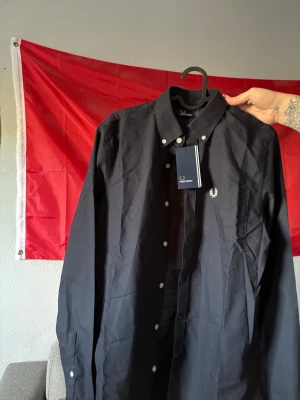 Svart skjorta från Fred Perry - Stilren svart skjorta från Fred Perry med klassisk krage och vita knappar. Skjortan har lång ärm och en liten broderad logga på bröstet. Perfekt för dig som gillar en clean och tidlös look.