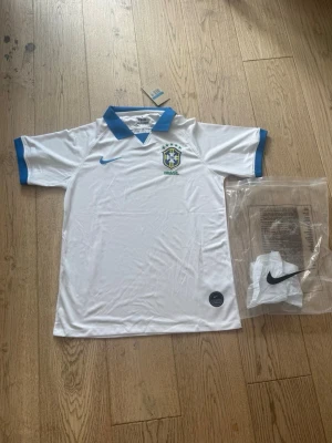 Vit Brasilien fotbollströja Nike - Vit Brasilien fotbollströja från Nike med blå detaljer på krage och ärmslut. Tröjan har Brasilien-emblemet på bröstet och Nike-logga i blått. Tillverkad i lätt och ventilerande material, perfekt för träning eller match.