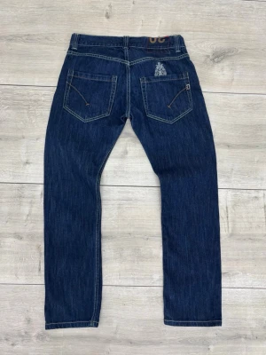 Mörkblå DONDUP jeans  - Säljer ett par mörkblå jeans från Dondup med raka ben och klassisk femficksmodell. Jeansen har kontrastsömmar och en broderad logga på ena bakfickan. Tillverkade i slitstark denim, perfekta för en clean och stilren look.