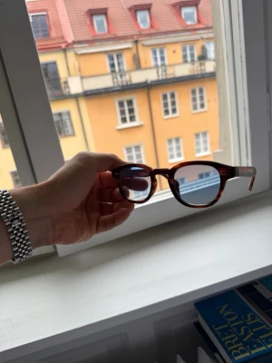 Solglasögon med blått glas oldmoney  - Snygga solglasögon med rundad form och brunmelerad båge. Glasen är blåtonade och ger en cool vibe. Perfekta för att lyfta din stil under soliga dagar.