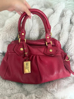 Röd Marc Jacobs handväska i läder - Säljer en snygg röd handväska från Marc Jacobs i mjukt läder. Väskan har dubbla handtag, guldfärgade metalldetaljer och en dekorativ metallbricka framtill. Rymlig modell med fina veck och detaljerade sömmar. Köpt här på plick, använt mycket men har enbart några små defekter 