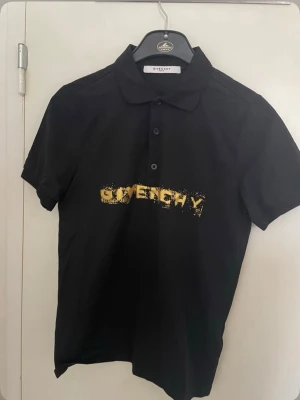 Svart Givenchy skjorta med gul logga - Svart skjorta från Givenchy med klassisk krage och knappar framtill. Tryckt gul Givenchy-logga på bröstet med en cool, sliten effekt. Skjortan har korta ärmar och är tillverkad i mjuk bomull som känns skön mot huden.