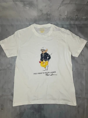 Vit Polo Bear t-shirt från Ralph Lauren - Vit t-shirt från Ralph Lauren med Polo Bear-motiv på framsidan. Klassisk rund hals och korta ärmar. Trycket visar en björn med cool outfit och texten 'Polo Bear by Ralph Lauren'. Perfekt för dig som gillar ikoniska prints och streetstyle.