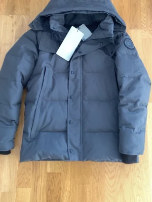 Grå dunjacka från Canada Goose XS - Snygg grå dunjacka från Canada Goose i modellen Expedition Parka Fusion. Jackan har huva, stora fickor och klassisk patch på ärmen. Perfekt för kalla vinterdagar med sin tjocka vaddering och stilrena design.