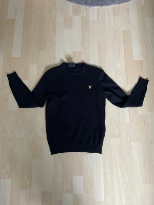 Svart stickad tröja från Lyle & Scott - Svart stickad tröja från Lyle & Scott med rund halsringning och den klassiska gula örnloggan broderad på bröstet. Tröjan har långa ärmar och ribbade muddar vid ärmslut och nederkant. Perfekt för en stilren look.