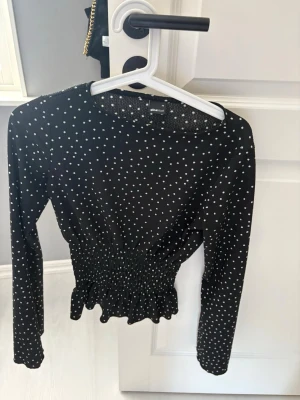 Svart prickig topp från Ginatricot - Svart långärmad topp från Ginatricot med vita prickar. Toppen har smockad midja och volangkant nedtill som ger en snygg siluett. Perfekt att styla med jeans eller kjol för en trendig look.