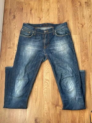 Blå jeans från Nudie Jeans - Säljer ett par klassiska blå jeans från Nudie Jeans med snygga slitningar och kontrastsömmar. Använda men har varit reparerade på skrädderi. Storlek 33/34 