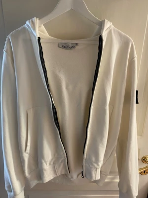 Vit hoodie från Stone Island - Säljer en vit hoodie från Stone Island med dragkedja framtill och två fickor. Klassisk svart patch med logga på vänster ärm. Hoodien har huva och ribbade muddar. Tillverkad i mjuk bomull för skön känsla och stilren look.