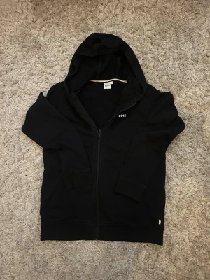 Svart zip hoodie från BOSS - Snygg svart hoodie från BOSS med dragkedja framtill och huva. Diskret BOSS-logga på bröstet och ribbade muddar vid ärmar och nederkant. Perfekt för en chill och stilren look. Tillverkad i mjukt material för extra komfort.