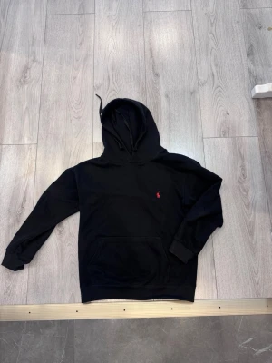 Svart hoodie från Ralph Lauren - Svart hoodie från Ralph Lauren med klassisk huva och känguruficka. Broderad röd logga på bröstet ger en stilren touch. Mjuk bomullskänsla och ribbade muddar vid ärmslut och nederkant. Perfekt för en avslappnad och snygg look.